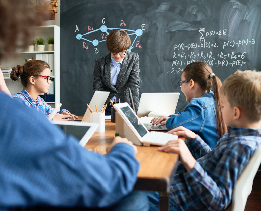 students around a desk with ipads and a chalkboard in the background showing equations, with another student standing just ahead of it with a smile